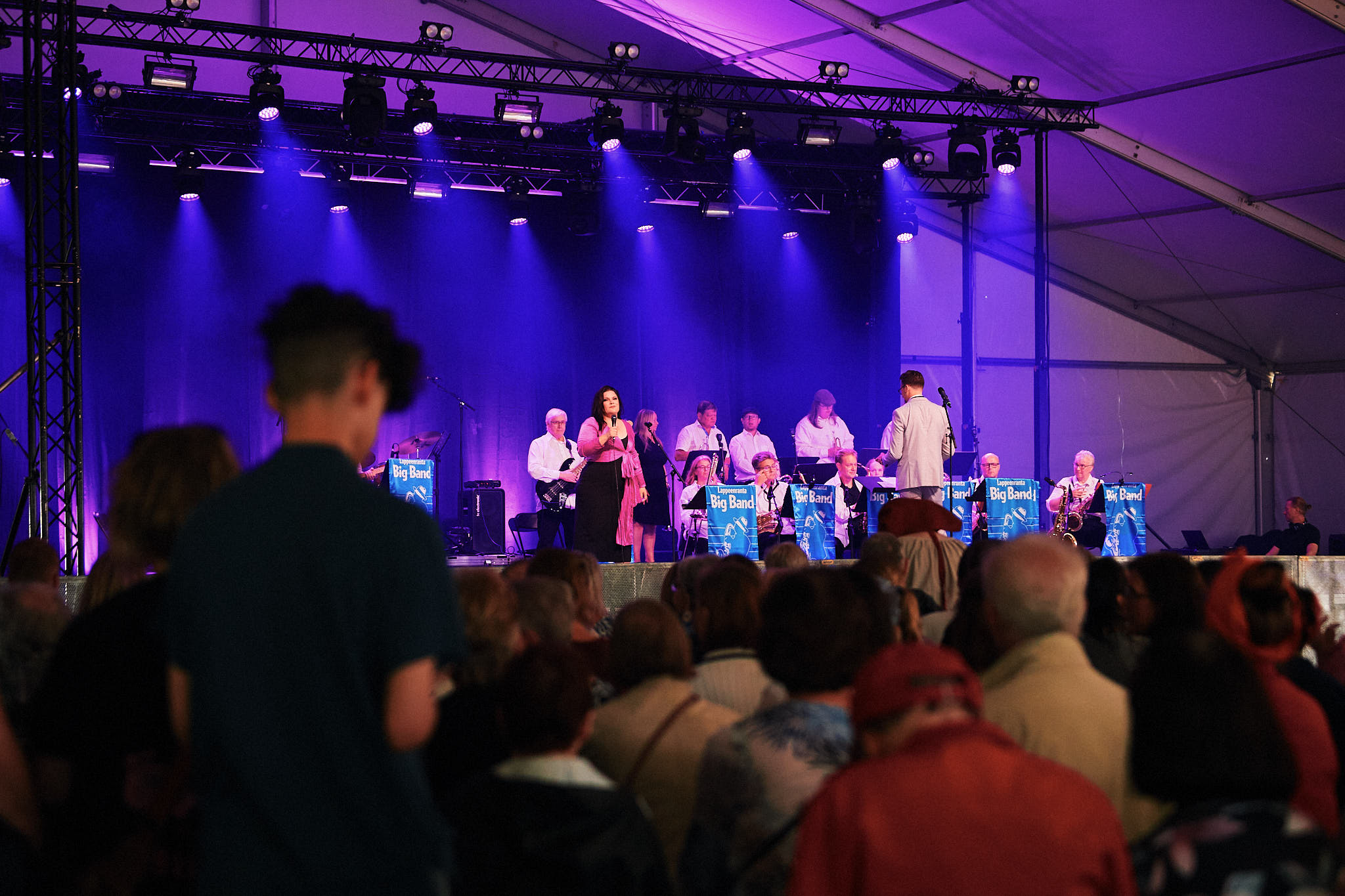 Ihmiset kuuntelevat teltassa musiikkiesitystä. Lavalla seisoo nainen mikrofoni kädessä, taustalla on big band orkesteri.